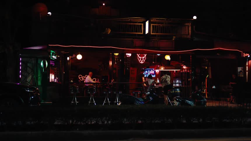 An open air roadside bar glows with neon and ambient lighting at night with several patrons seated on stools watching a screen and socializing