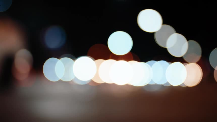 Cars drive through the city at night with headlights that are blurry and unrealistically large