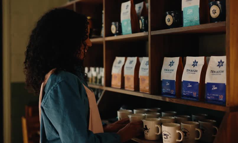 A young woman arranges coffee mugs on a shelf surrounded by colorful coffee bags in a welllit shop