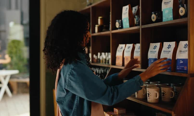 A woman wearing a denim apron organizes shelves filled with coffee bags mugs and decor in a brightly lit shop