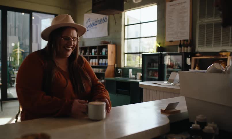 A woman wearing a hat and an orange sweater receives a cup across a counter in a cozy caf with large windows and a menu board on the wall