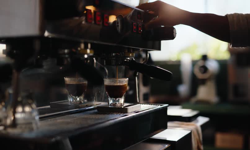 A persons arm seen using a coffee maker to serve a glass of hot coffee in a glass inside a coffee shop at midday