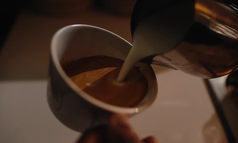 A person pours coffee in a cup and then uses cream to make a design