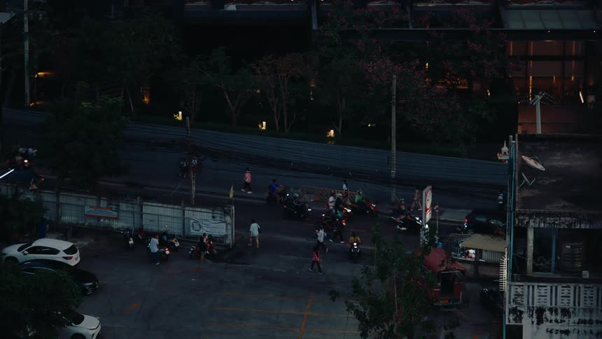 At dusk a parking lot bustles with activity as people gather around parked cars and motorcycles near a building