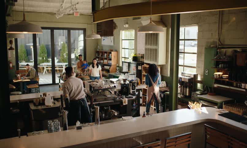 A cafe with patrons enjoying drinks at tables and baristas working behind a counter under warm industrial lighting