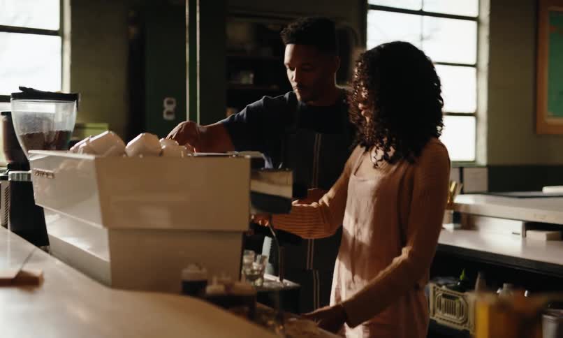 Two individuals work in a coffee shop one operating an espresso machine and the other observing nearby