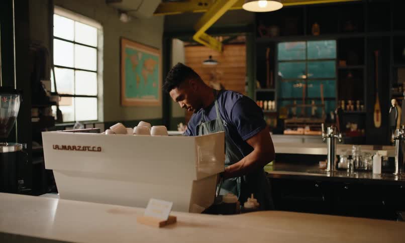 A barista prepares a drink behind a coffee shop counter adorned with pastries and coffee equipment with large windows and a map on the wall