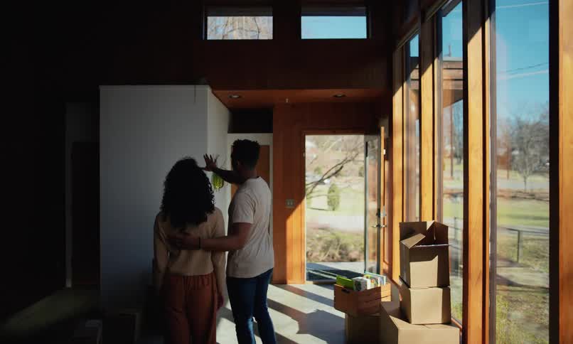 A man and a woman are inside a house with glass walls and looking at a white wall and talking about