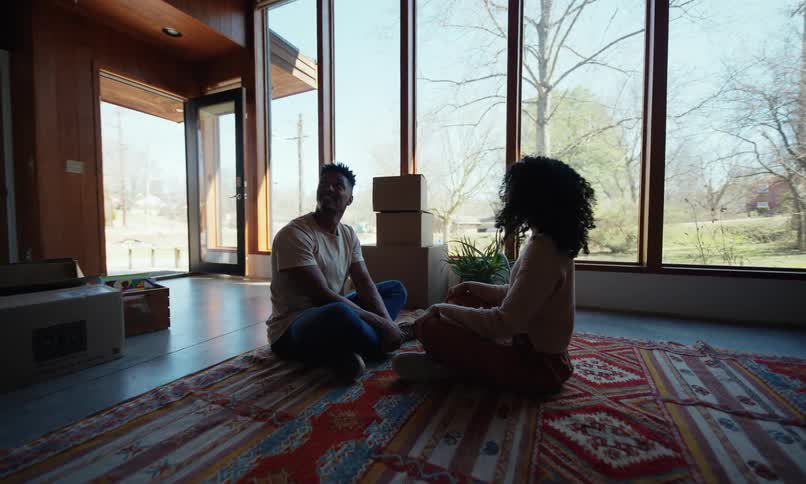 Two people sit comfortably on a colorful patterned rug surrounded by unpacked boxes in a spacious sunlit room with floortoceiling windows