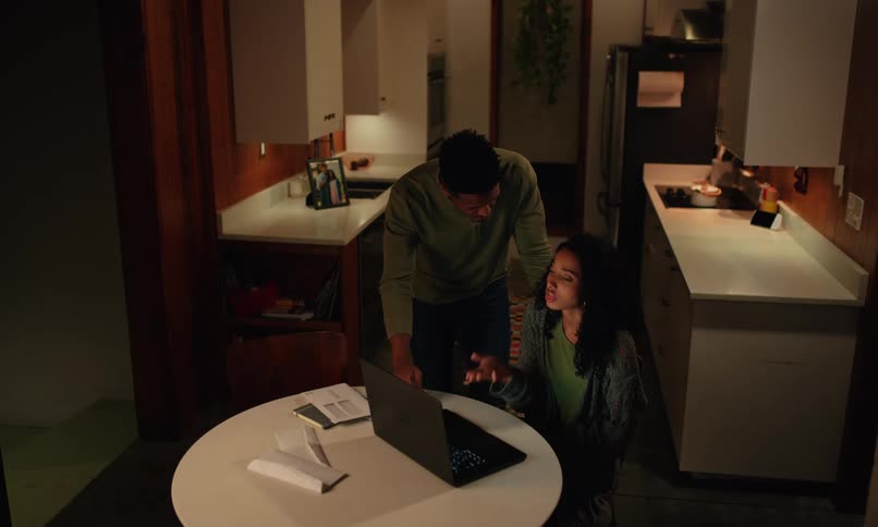 A black woman argues with a man while she sits in the kitchen and looks at her laptop