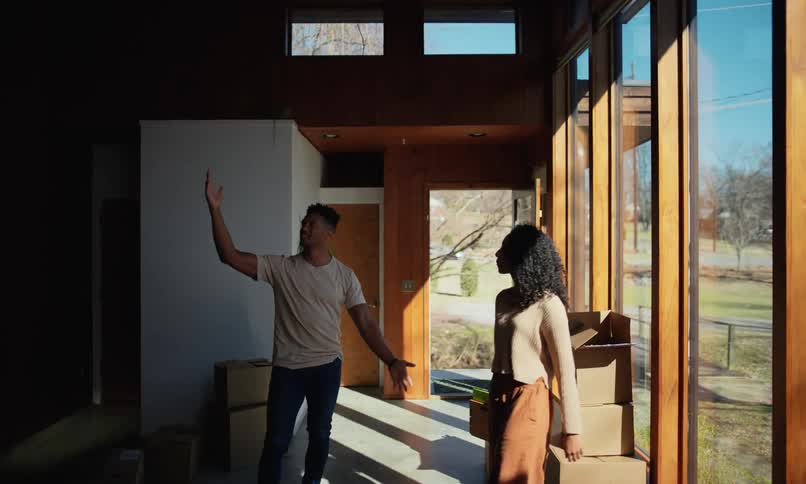 A young couple embrace in a sunlit room with large windows surrounded by moving boxes and furniture