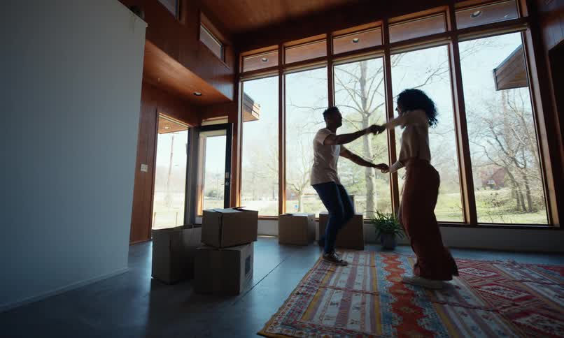 Couple dances in their new home near floor to ceiling windows showing leafless trees during the daytime