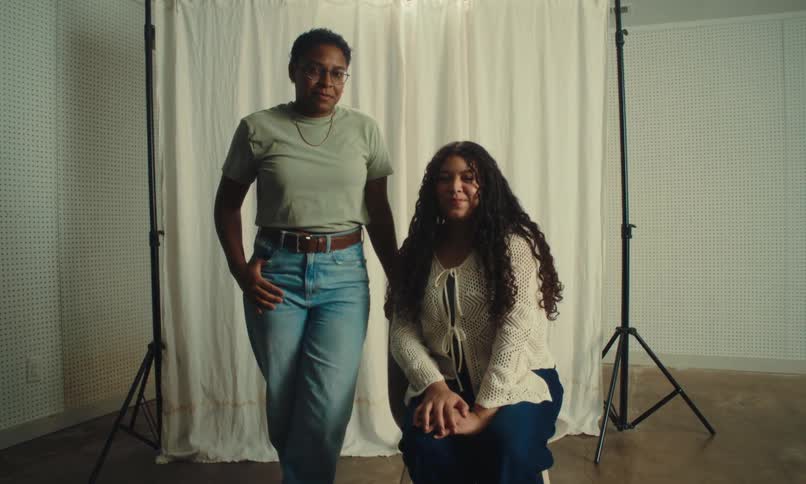 Two women are positioned in front of a white curtain backdrop with one standing and the other sitting on a stool in a room equipped with photography lighting equipment