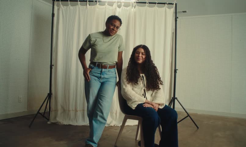 Two individuals are positioned in front of a white curtain backdrop with one standing and the other sitting on a chair in a room equipped with photography lighting equipment