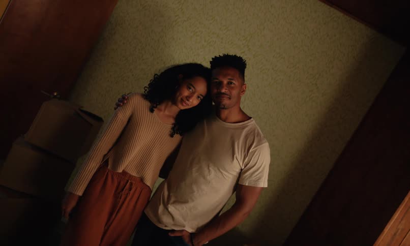 A man and a woman are hugging and staring straight ahead with a subtle smile in an indoor setting