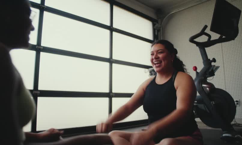 Two curvy women, one white and the other black, sitting on a yoga mat and giving each other a high five in the air both smiling and happy after a day of exercising. A stationary bike is behind them next to the wall