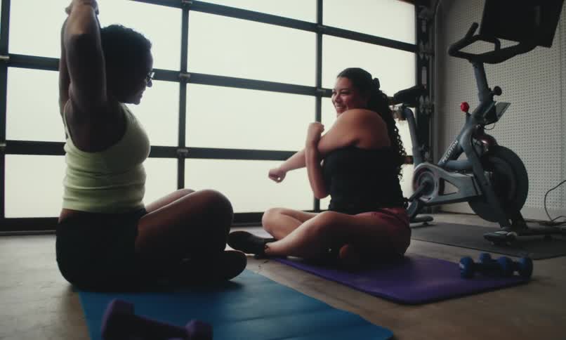 Two women are on yoga mats in a gym as they stretch