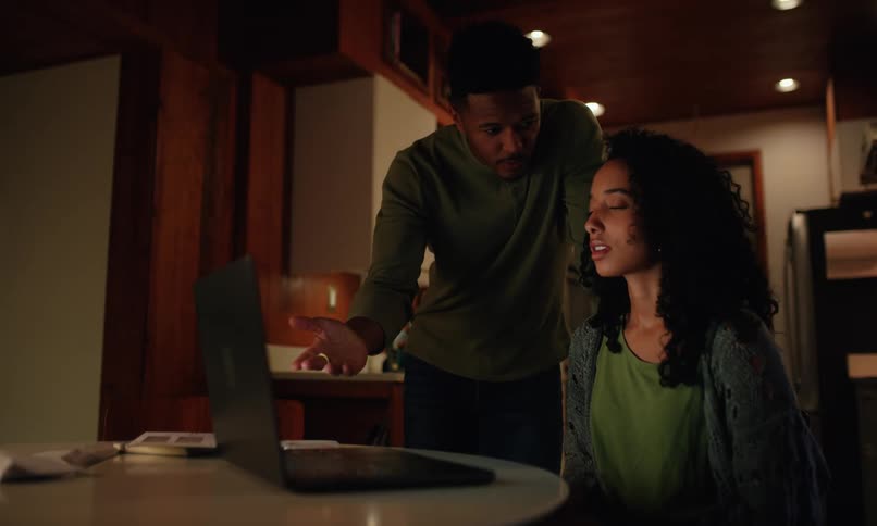 A couple are in a kitchen one standing and gesturing toward a laptop on a table while the other sits frustrated