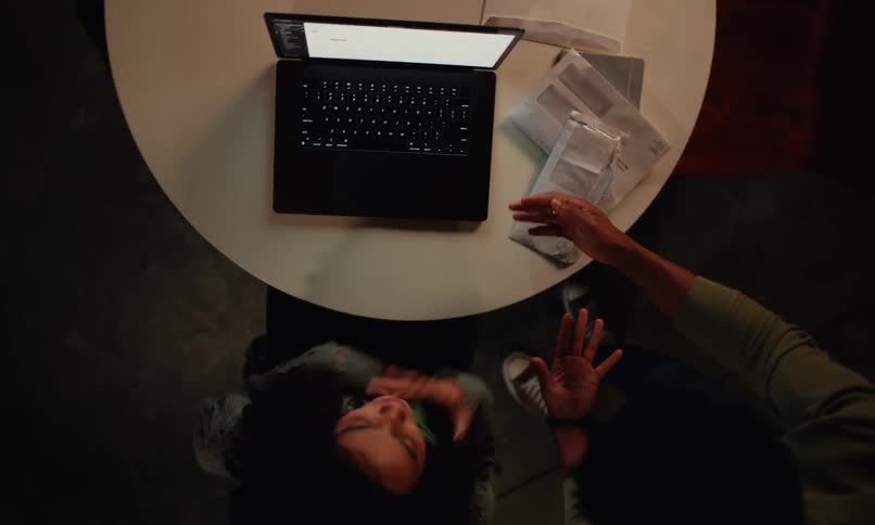 A black couple arguing in front of a computer placed on a white coffee table with the woman seated and the man standing beside her