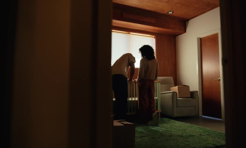 A young couple inside a room filled with moving boxes are talking to each other while facing a window examining a infant bed in front of them at midday