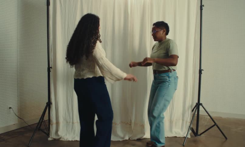 Two women dance near a white curtain backdrop in a bright minimal setting