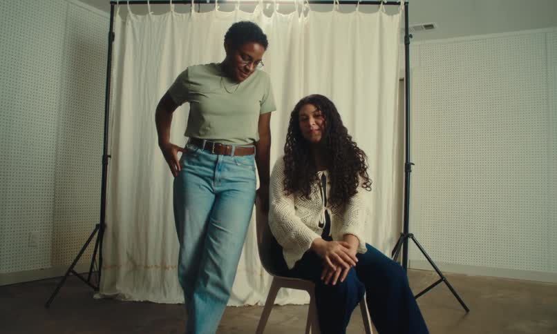 Two people are in front of a curtain one sitting in a chair and the other standing and leaning on the chair