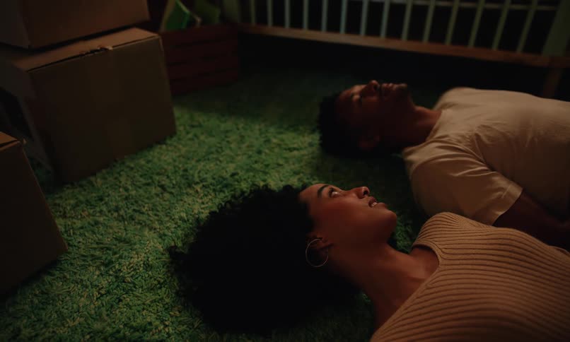 Two people lounge on a green carpet surrounded by moving boxes