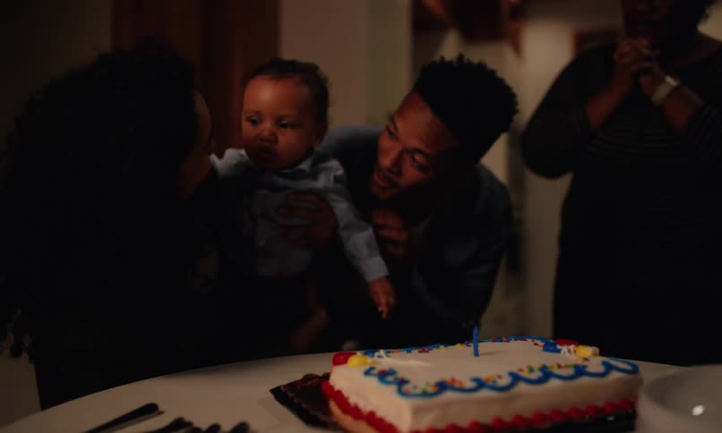 A man and a woman are happily celebrating with a child on their lap and blowing on a birthday cake that is on a table in front