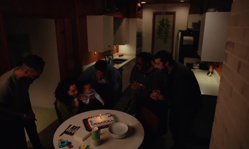 A family gathers in a cozy kitchen around a table adorned with a birthday cake toy truck and festive decorations to celebrate a babys birthday
