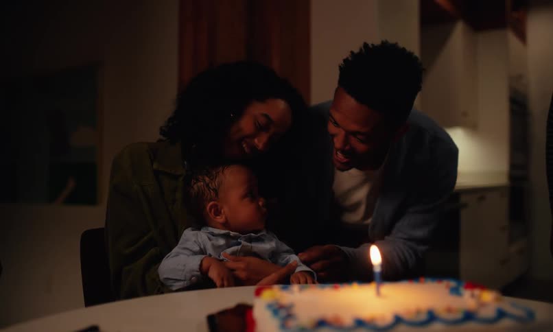 A father lights a birthday candle for his son who is being held by the mother as they both happily sing a birthday song for him
