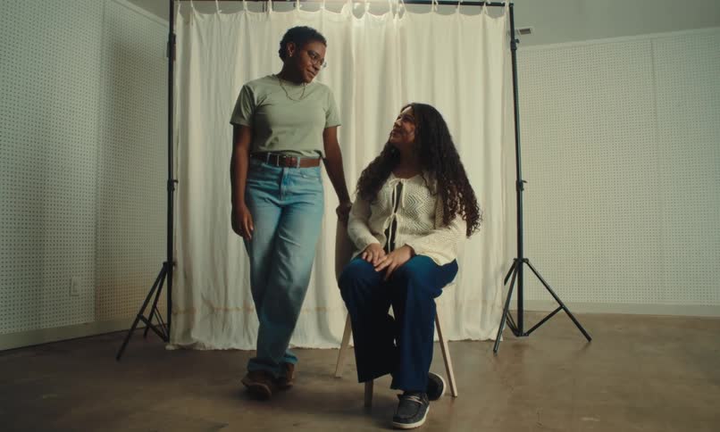 A couple of two women where one is standing and the other is sitting and the one who is standing removes the hair that is in the eye of the one who is sitting