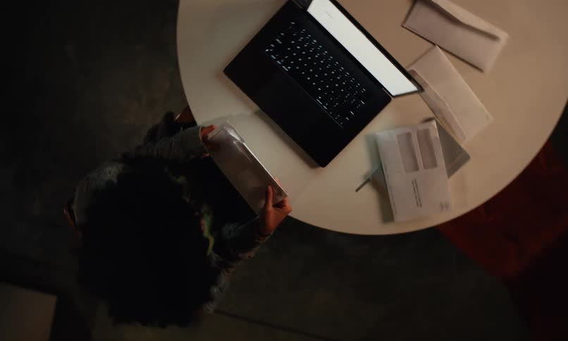 A worried woman sits at a table while opening mail and looking at her laptop
