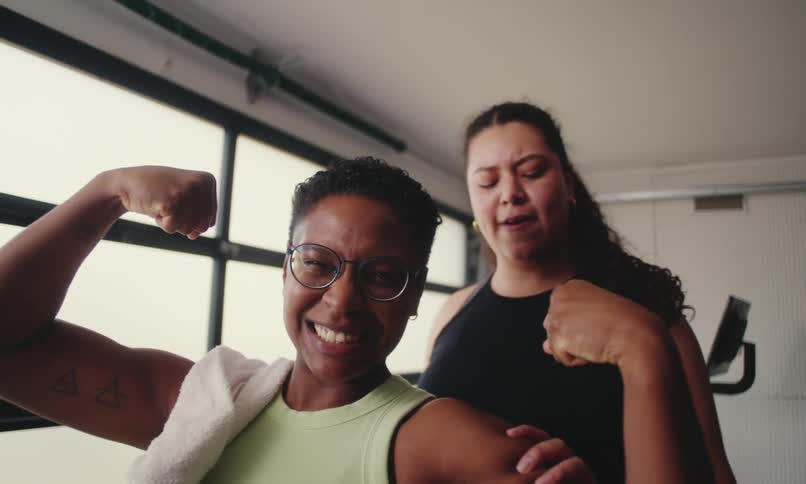 Two women flex their arm muscles in a welllit room with large windows and minimal furnishings