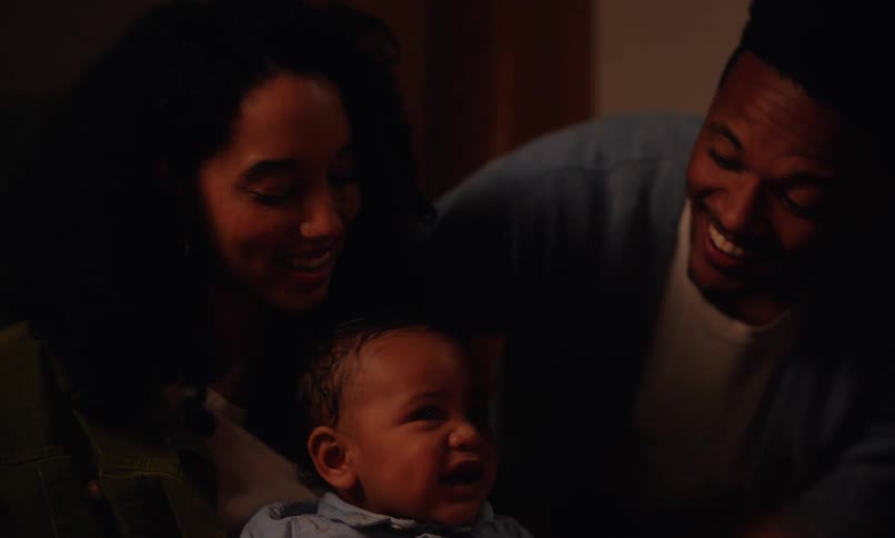A man and a woman are happy and holding a little baby in their arms while they are looking at a birthday cake that is in front with a lit candle