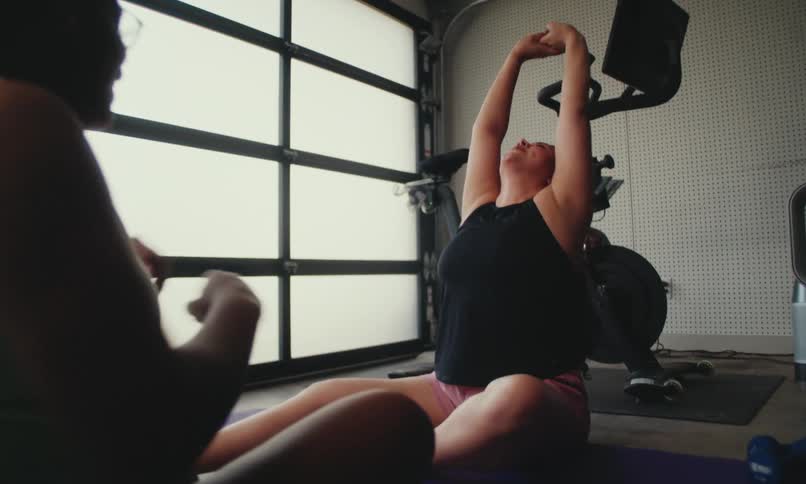 Two women sitting on yoga mats in a gym at midday facing each other as they stretch their arms sideways