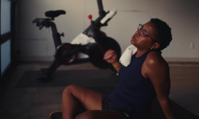 A black woman sitting on the floor on a yoga mat inside a gym wearing a tank top and shorts looks exhausted and tired as she wipes sweat from her face with a napkin