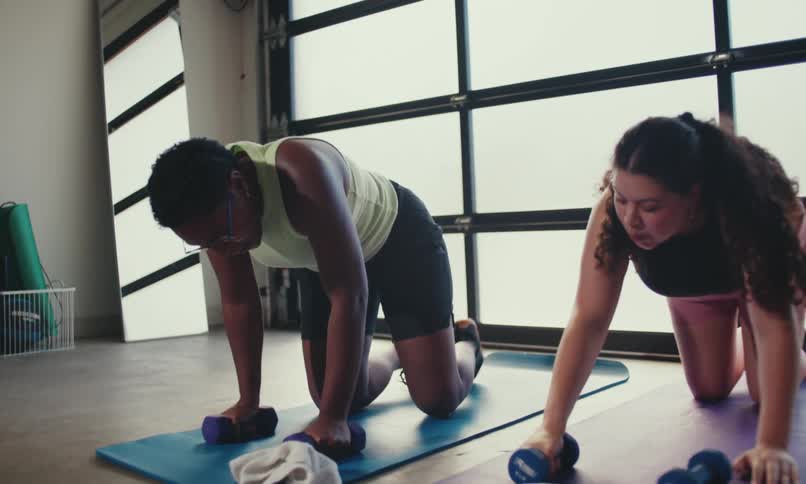 Two people exercise on yoga mats in a bright room performing plank dumbbell rows