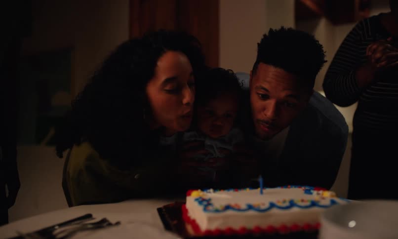 Parents with their small child on their lap and other people around blowing out a candle together with their child at a birthday party