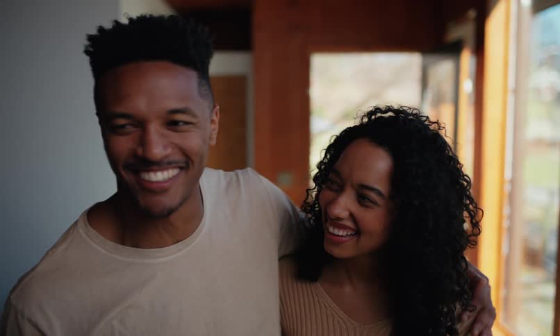 A happy young couple smiles and gestures while talking looking around their newly movedinto home with large windows and minimal furnishings