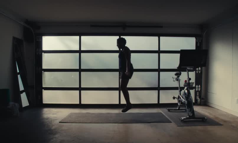 A woman jumps rope on a mat in a room with a large window and an exercise bike nearby