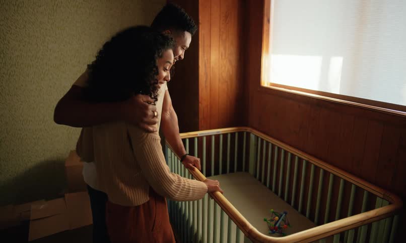 A couple embraces tenderly within a warmly lit slightly rustic apartment setting with a baby crib visible in the background
