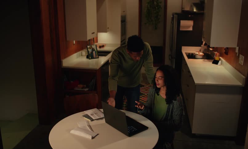 A woman working on a laptop while seated at a coffee table in the kitchen looks worried as a man standing nearby approaches her and they argue about what she is working on on the laptop at night