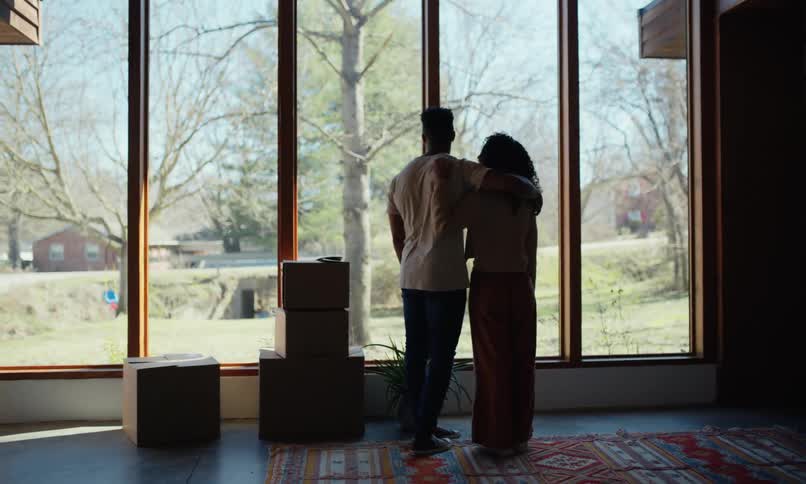 Young couple standing in living room looking out large window to their new front yard