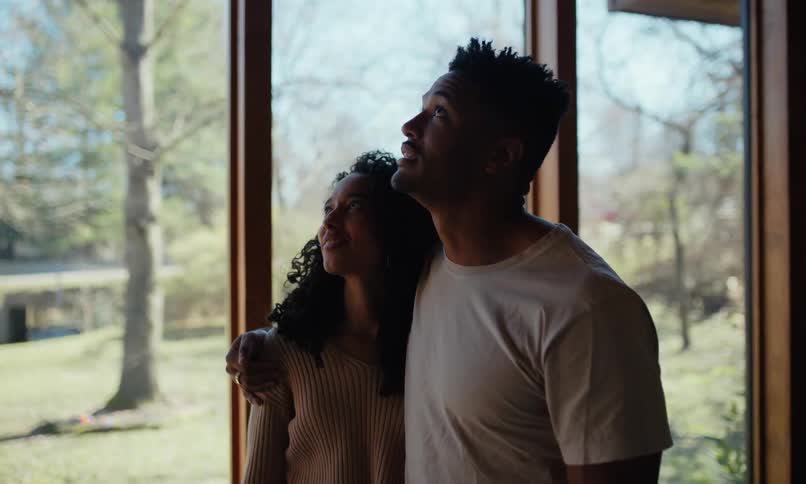 A young couple inspecting the house theyre moving into discussing it as moving boxes lie scattered on the floor they stand by the window with grass and trees visible outside during the day