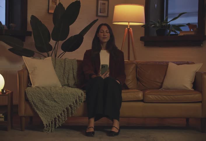 Woman with eyes closed sitting on living room sofa holding smartphone and taking deep breaths