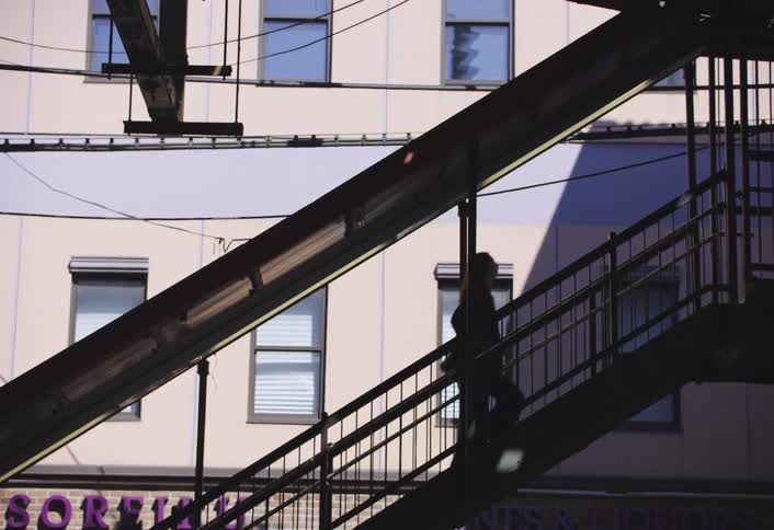 A young woman is seen climbing a staircase leading to an elevated train station on a sunny day with an apartment building situated right beside it