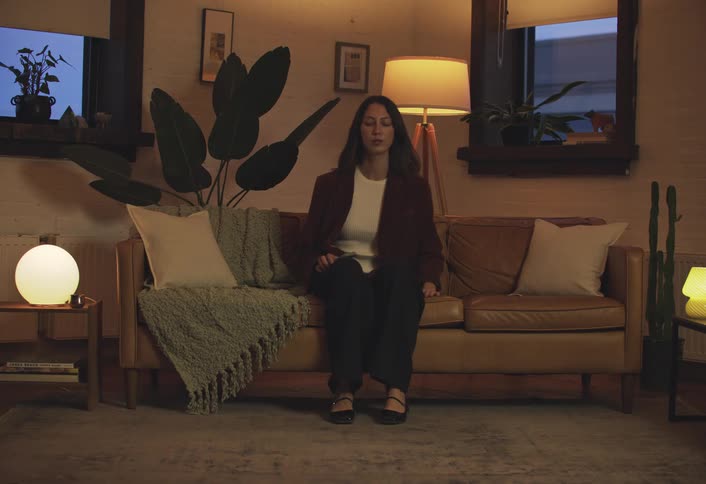 A woman walks calmly into the softly lit room and sits on the couch settling into a quiet reflective moment
