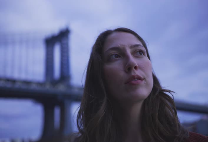 The woman is standing by a riverbank with a bridge visible in the background smiling as she looks down at her iphone
