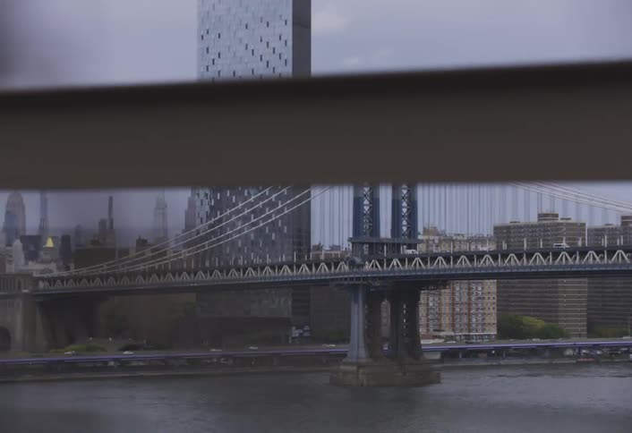 A wide bridge spans a flowing river amidst a modern cityscape featuring a reflective skyscraper and urban buildings viewed from a moving vehicle