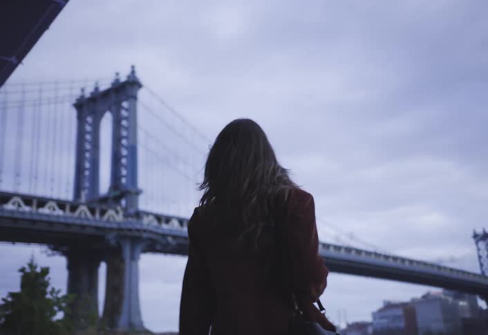 A woman walks towards the large bridge on a cloudy day